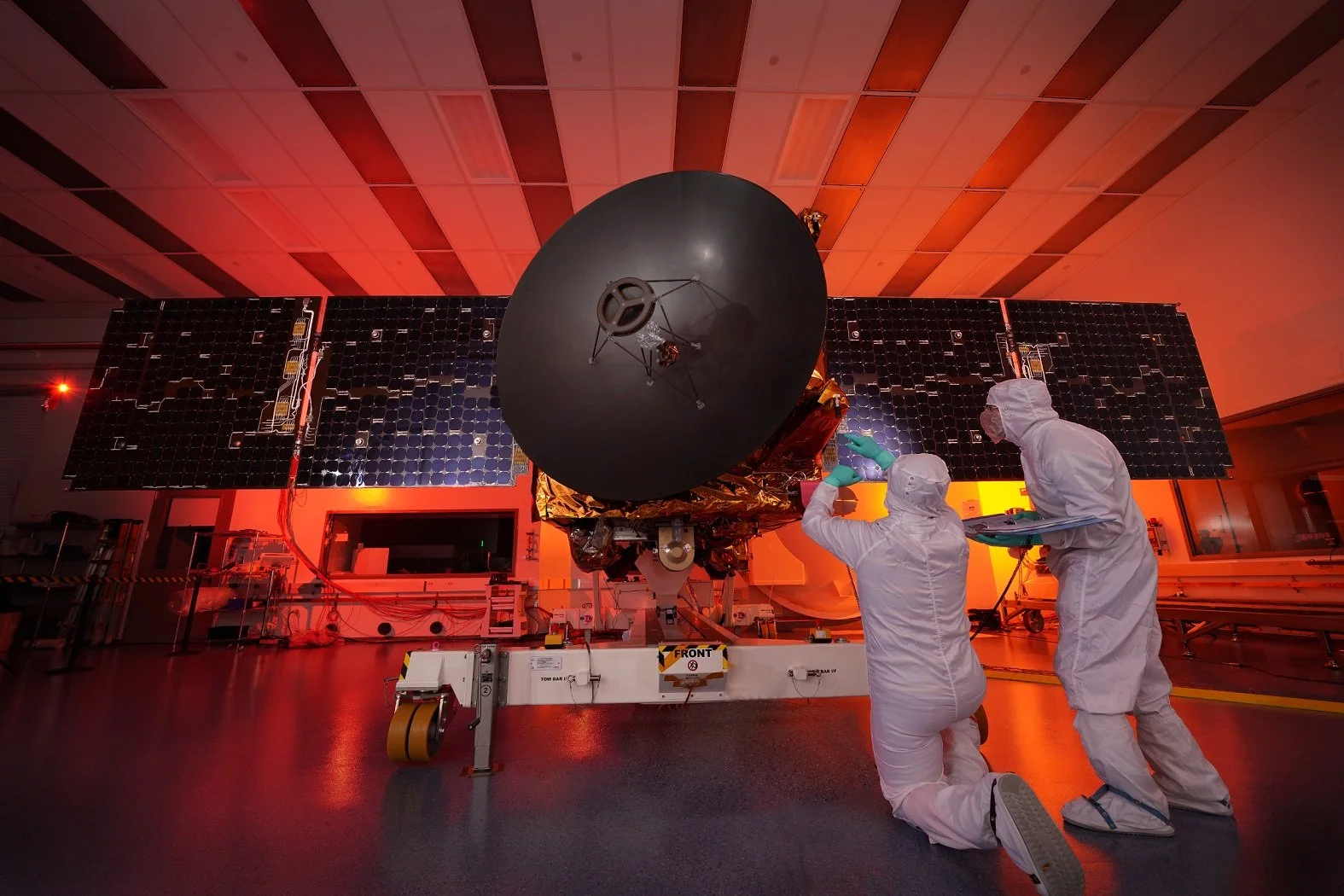 Two scientists in white protective suits working on a satellite or spacecraft inside a cleanroom with a red lighting ambiance.