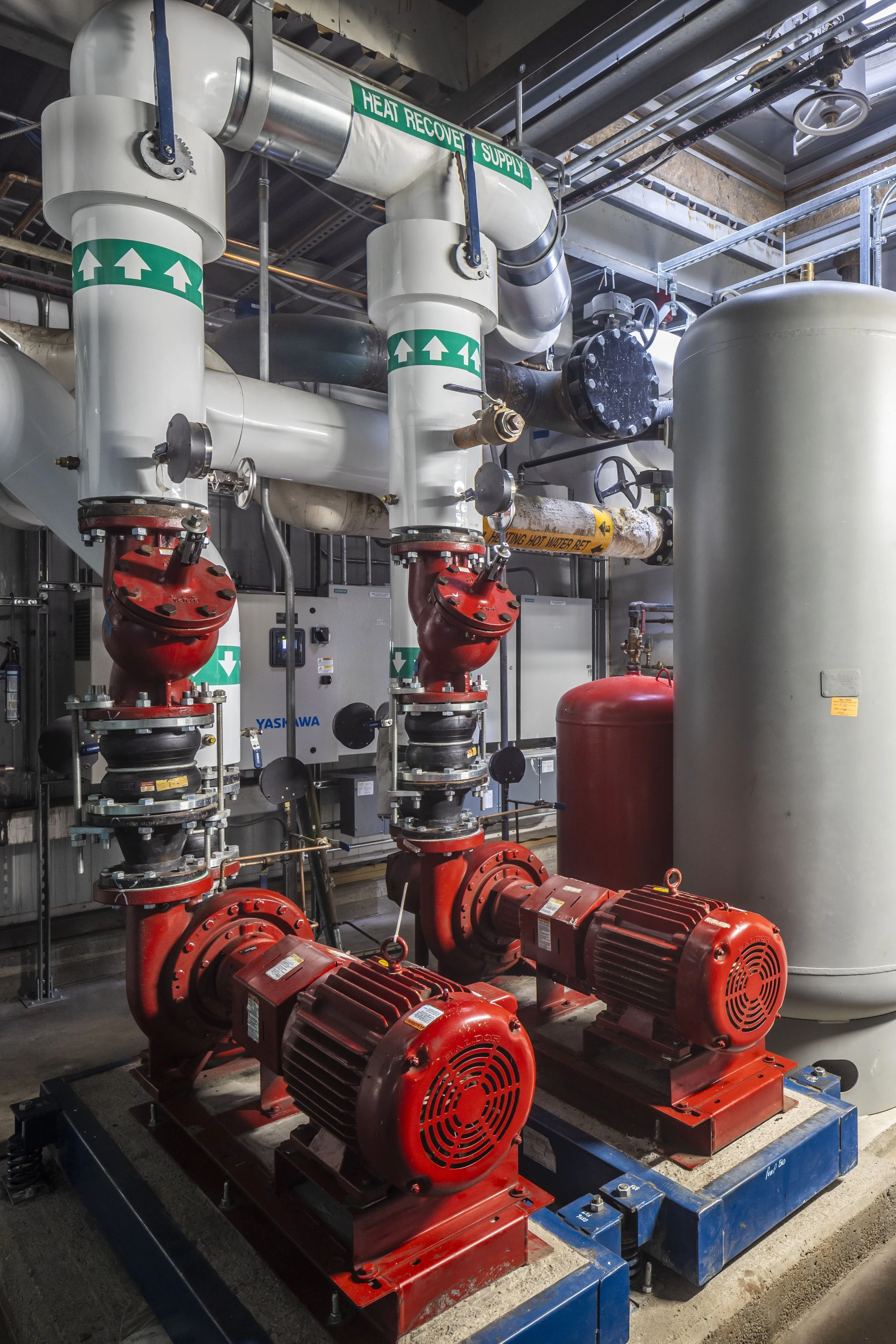 Industrial equipment with two large red motors, large white pipes labeled 'Heat Recovery Supply,' and a large gray tank in the background.