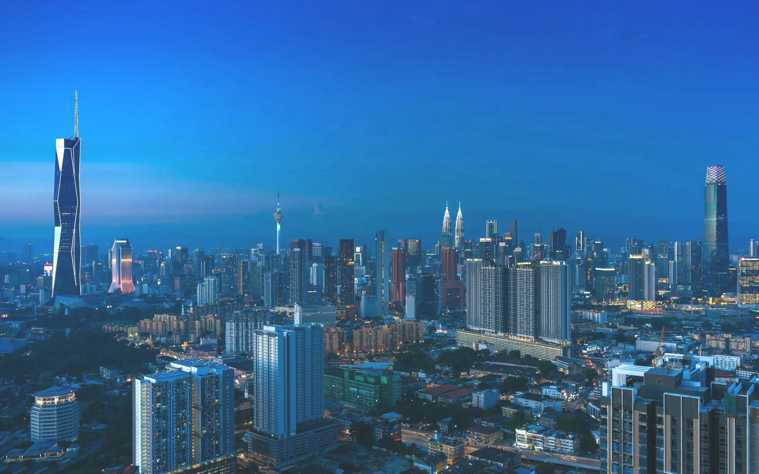 City skyline at dusk with tall skyscrapers, including the iconic Petronas Towers and Kuala Lumpur Tower, with a blue sky background.