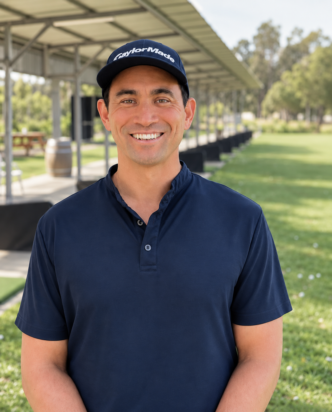 A man wearing a navy blue TaylorMade cap and matching polo shirt smiling outdoors in a park with a covered area and green grass in the background.