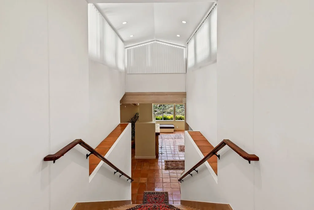 Interior view of a bright home with stairs leading down to a tiled living space with large windows and greenery outside.