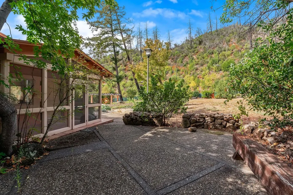 A backyard patio surrounded by trees and shrubs, with a view of a grassy field and a hillside in the background, under a partly cloudy sky.