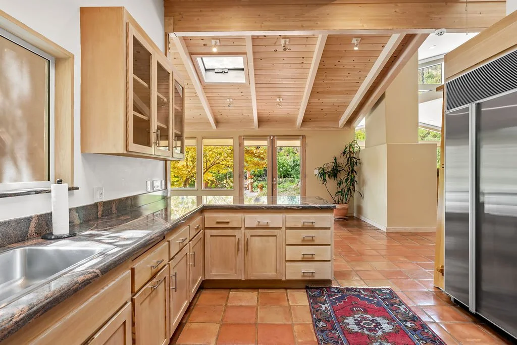 Kitchen with wooden cabinets, granite countertop, stainless steel sink, tile floor, large windows, potted plant, and a rug.