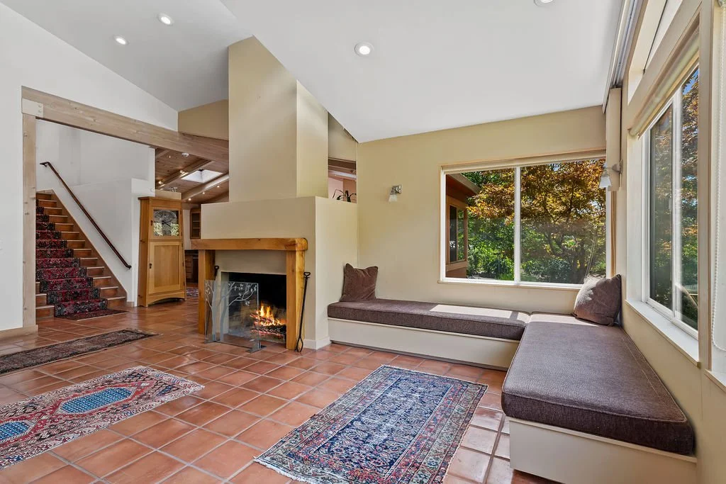 Living room with a fireplace, built-in window seat with cushions, large windows showing trees outside, terracotta tiled floor, and a staircase leading to upper level.