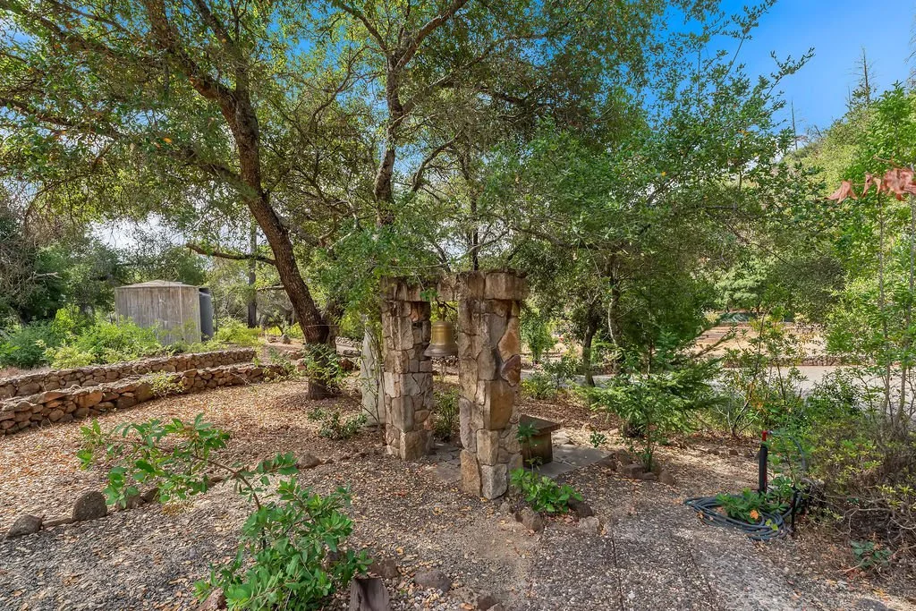 A rustic outdoor garden area with a stone archway and hanging bell surrounded by trees and shrubs, with a stone wall and shed in the background.