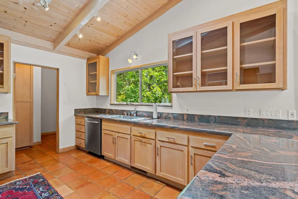 A kitchen with light wood cabinets, granite countertops, and a window overlooking green trees. The ceiling is wooden with recessed lighting, and the floor has terracotta tiles.