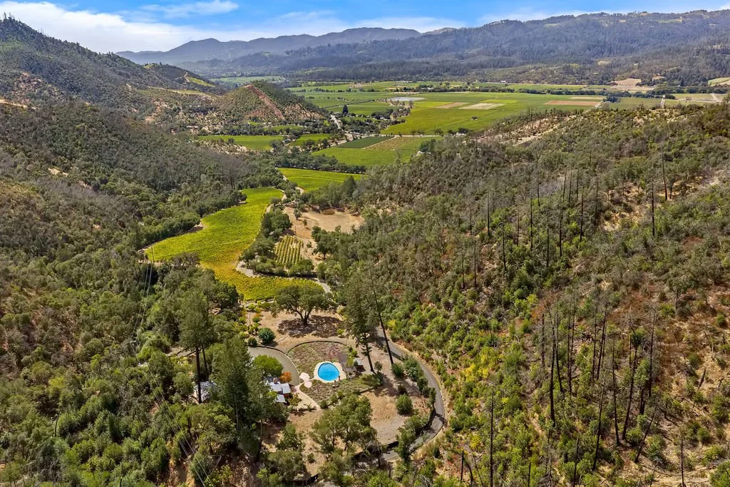 A vast valley surrounded by green hills and mountains, with cultivated fields and a small residential area featuring a swimming pool.
