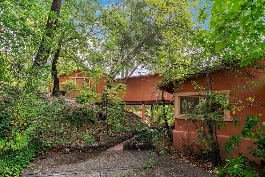A house with a pink stucco exterior surrounded by lush green trees and bushes, with a stone-lined pathway leading up to it.