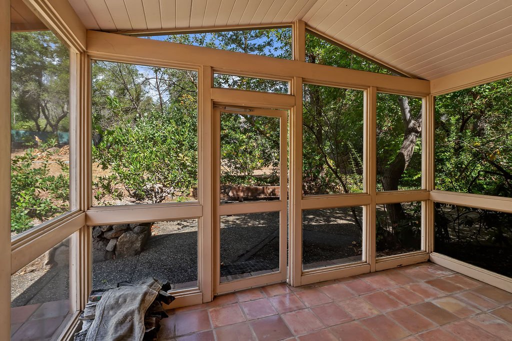 Sunroom with large windows overlooking trees and bushes outside, tiled floor, and a blanket or cloth on the floor in the corner.