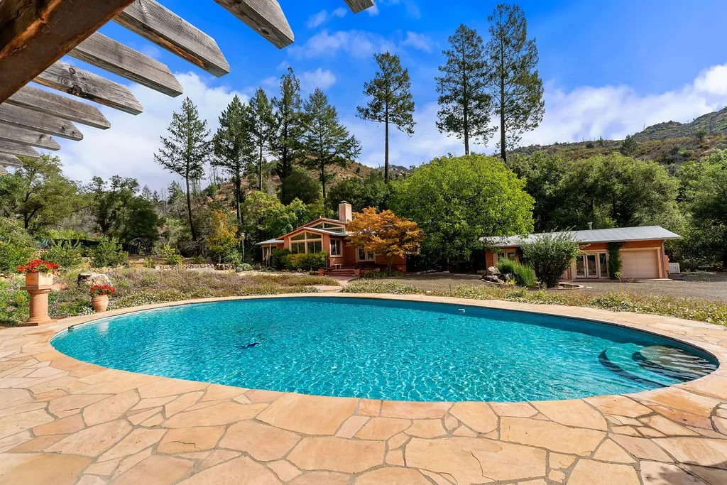 A backyard with a large swimming pool surrounded by a stone patio, trees, and a house and garage in the background under a partly cloudy sky.
