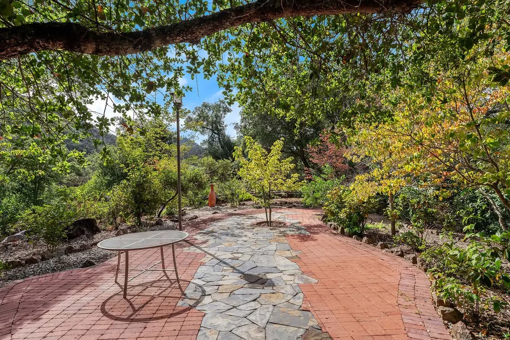 A winding garden pathway with a mix of red brick and natural stone tiles, surrounded by lush green trees and shrubs, under a canopy of overhanging branches.