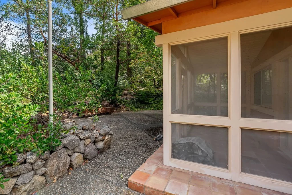 A corner of a house with screened-in porch, surrounded by a gravel pathway, rocks, greenery, and trees.