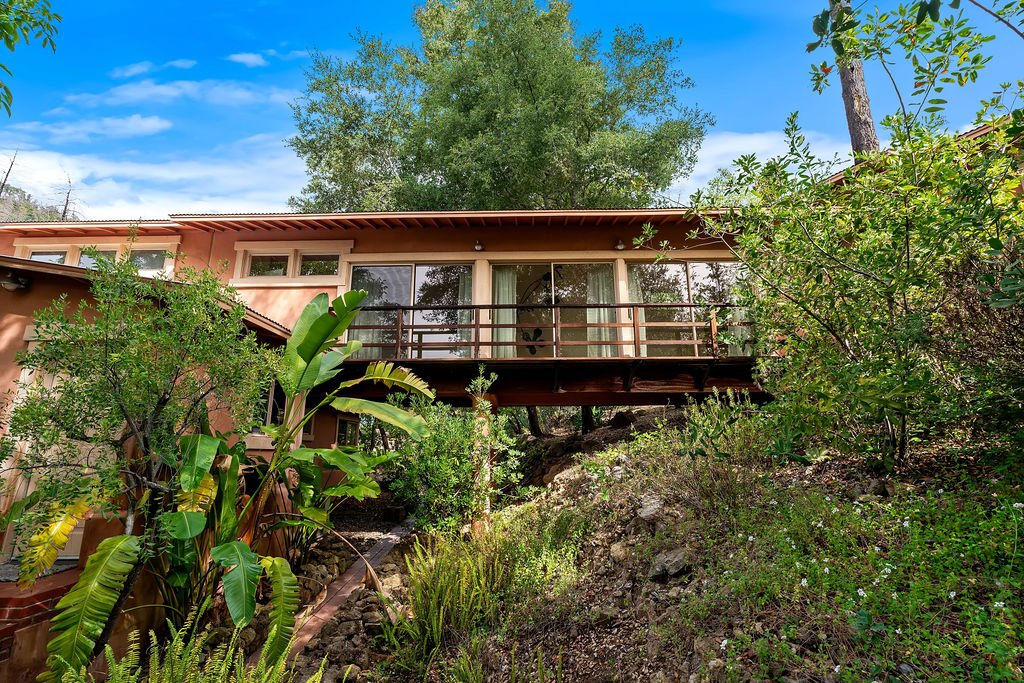 A house on a hillside with a glass sliding door, metal railing, and lush green plants and trees surrounding it.