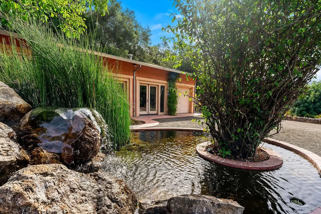 A backyard with a small pond surrounded by rocks and greenery, and a house with a garage in the background.