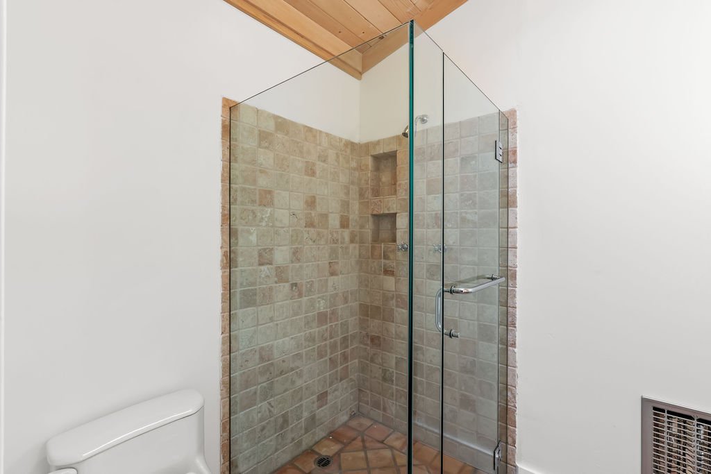 A walk-in shower with beige tile walls and a glass door, next to a white toilet, in a bathroom with a white wall and wooden ceiling.