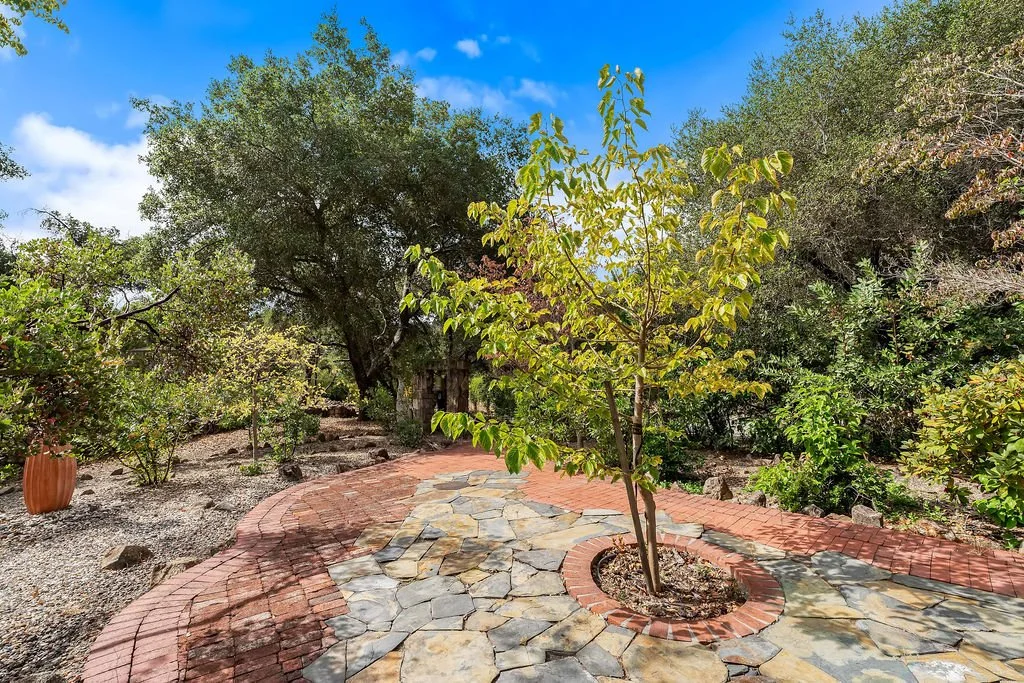A garden path with a meandering stone walkway, brick edging, and surrounding lush green trees and plants under a partly cloudy blue sky.