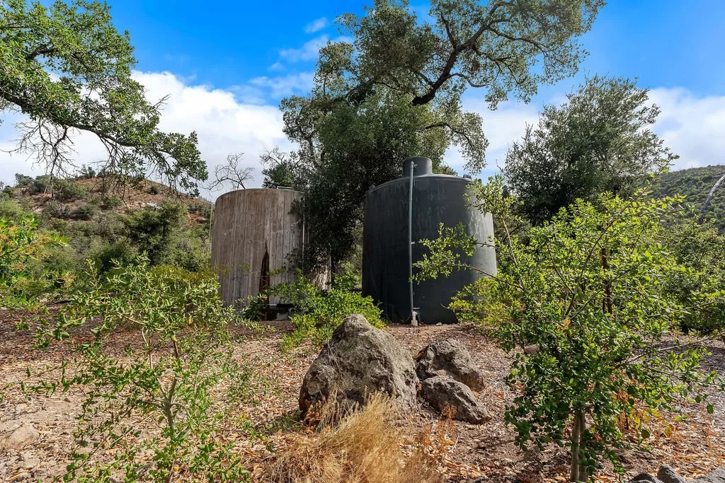 Two large water storage tanks, one wood-colored and the other black, are situated outdoors amidst bushes and trees with a hillside in the background and partly cloudy blue sky.