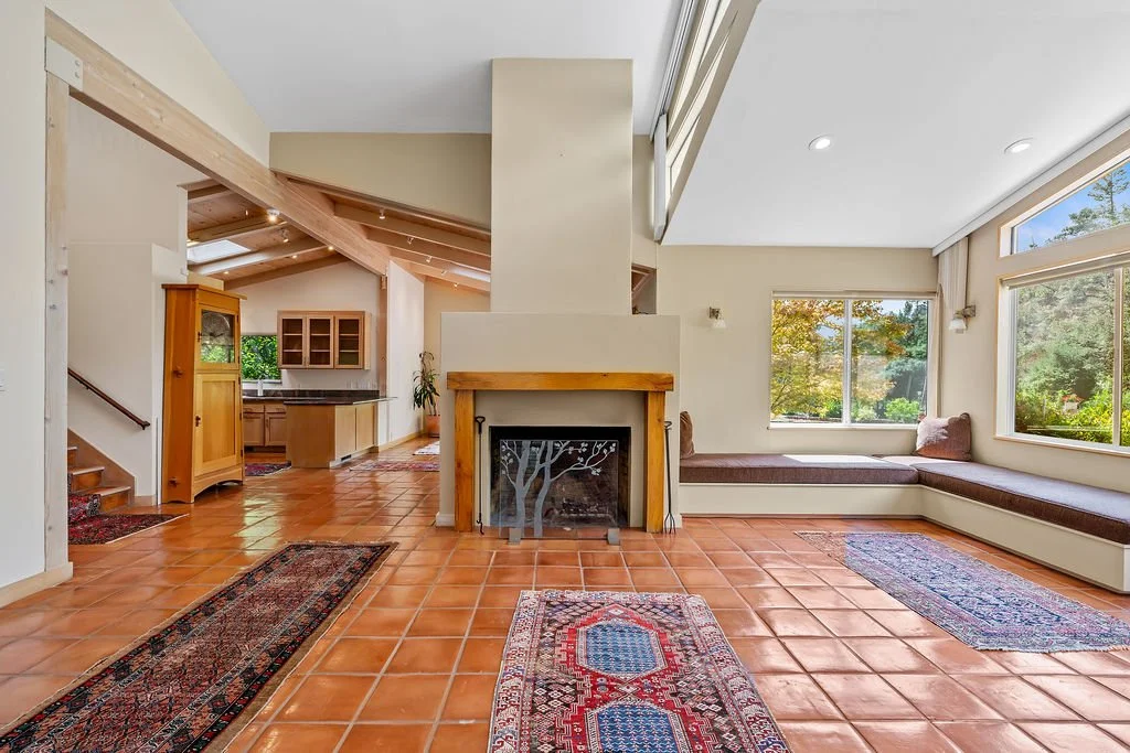 Spacious living room with large windows, built-in window seat, terracotta tile flooring, and a fireplace, connected to a kitchen area with wooden cabinets and sloped ceiling with skylights.