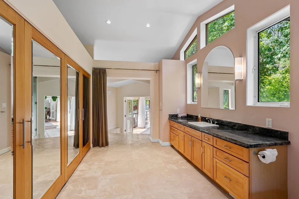 Bright laundry or utility room with wooden cabinets, black countertop, white sink, large windows with greenery outside, and mirrored sliding closet doors.
