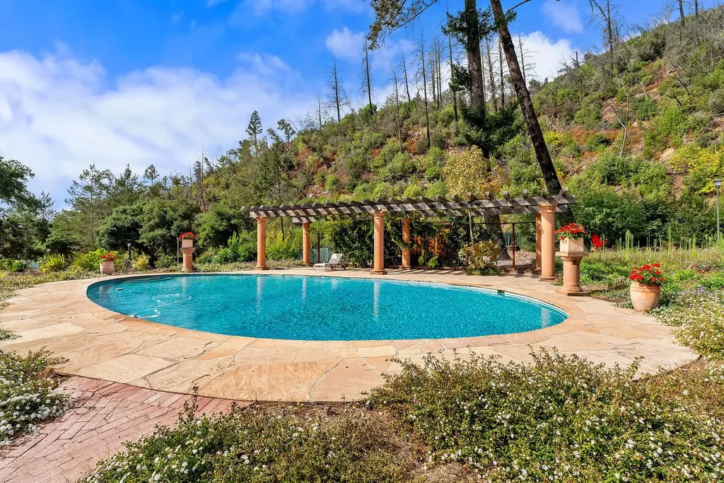A swimming pool surrounded by a stone patio with potted plants and a pergola in a natural setting with hills and trees.