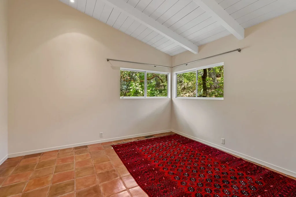 Empty room with white walls, tiled floor, and a red patterned rug. Two large windows showing green trees outside, and a white ceiling with exposed beams.