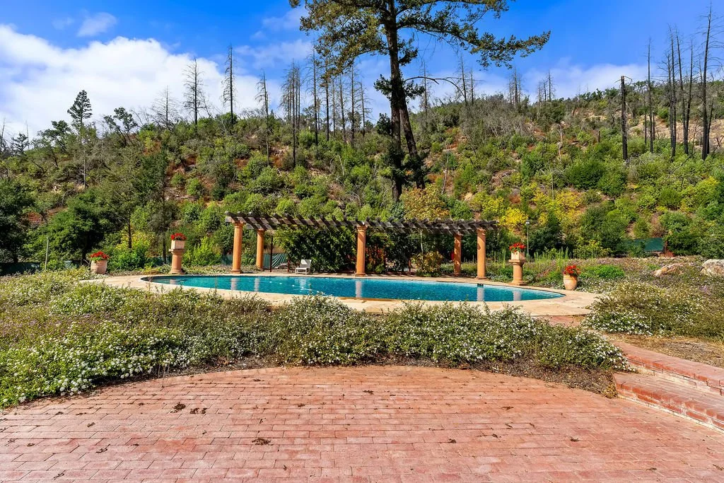 A swimming pool surrounded by greenery and flowers, with a hillside and trees in the background under a partly cloudy sky.