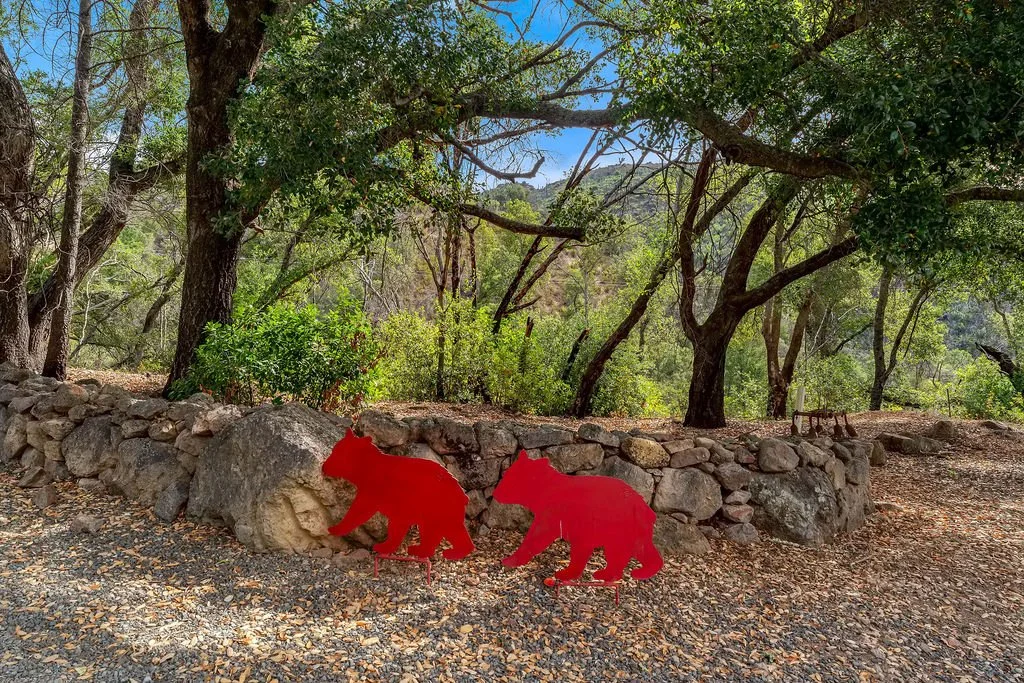 Two red metal bear sculptures positioned in front of a stone wall in a wooded outdoor area with trees and greenery.