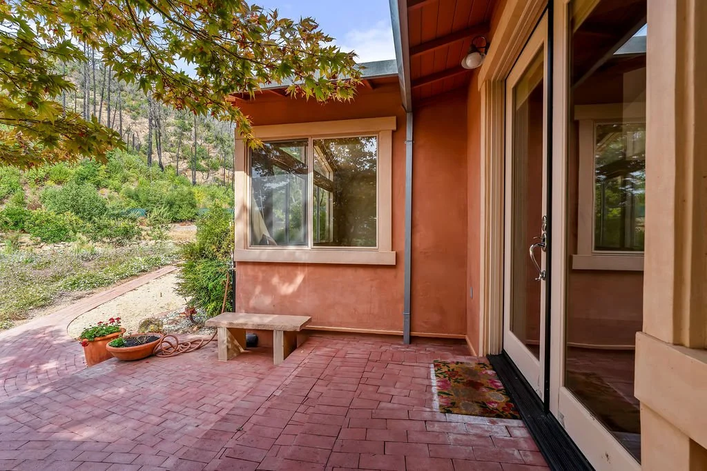 A small outdoor patio area with red brick flooring, potted plants, a wooden bench, and a sliding glass door leading inside a house. The house has ochre-colored stucco walls and large windows, with a backyard featuring greenery and trees in the backgr