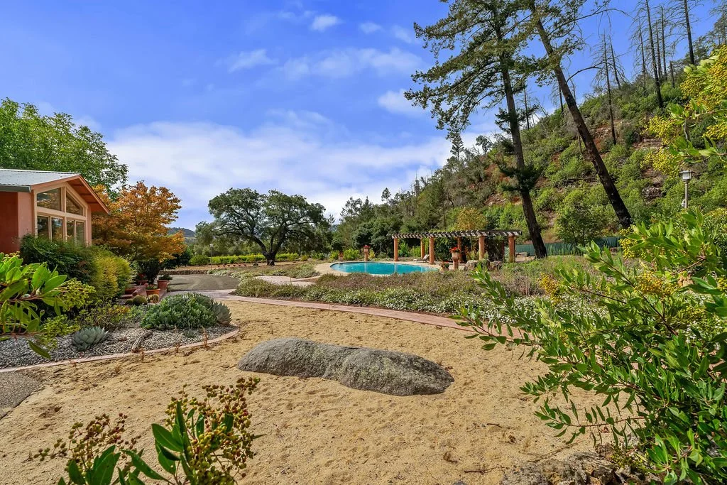 View of a landscaped backyard with a sandy area, large rocks, green plants, a swimming pool, porch with pergola, trees, and a mountain slope in the background under a partly cloudy blue sky.