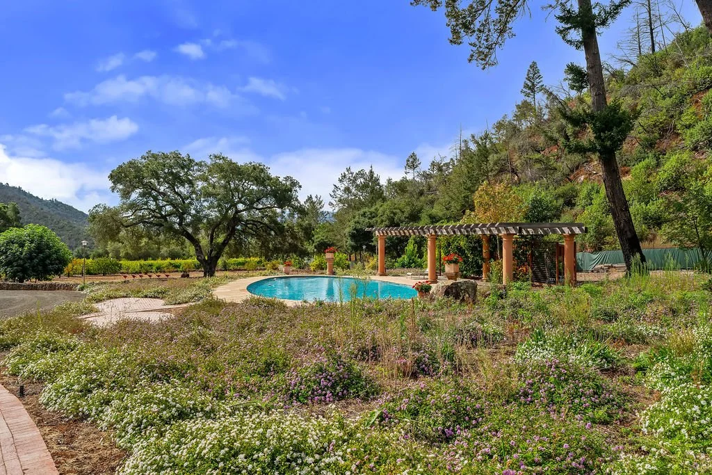 A scenic outdoor pool area surrounded by greenery and colorful flowers, with a large tree, a pergola, and hills in the background under a partly cloudy sky.