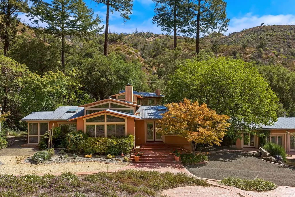 A modern house surrounded by trees and shrubbery, situated on a hillside with a gravel and paved driveway.