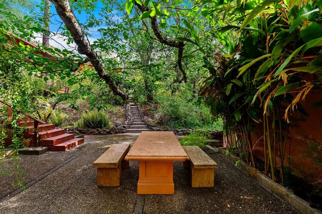 A peaceful outdoor garden with a stone table and benches, lush green trees, plants, and stairs leading up a hillside.