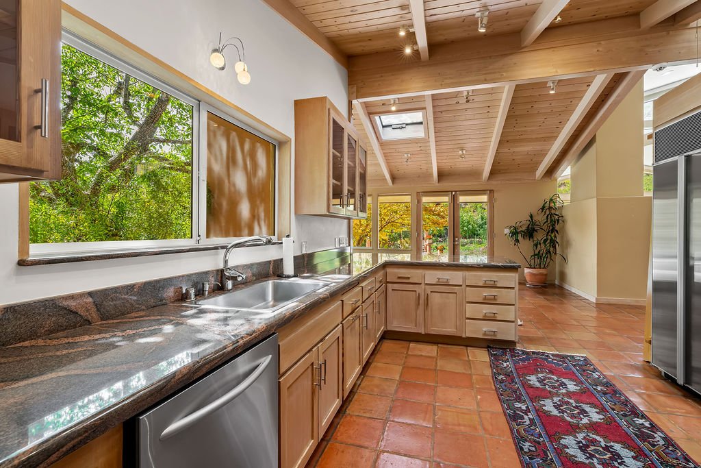A spacious kitchen with wooden cabinets, a granite countertop, and a stainless steel sink. Large windows show green trees outside. There is a patterned rug on the terracotta tile floor and a potted plant in the corner. The ceiling is wooden with expo