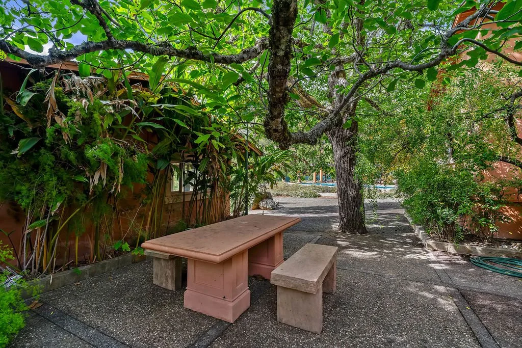 An outdoor garden patio with a stone table and matching bench, surrounded by lush green trees and plants, with a gravel pathway and a wooden house or shed in the background.
