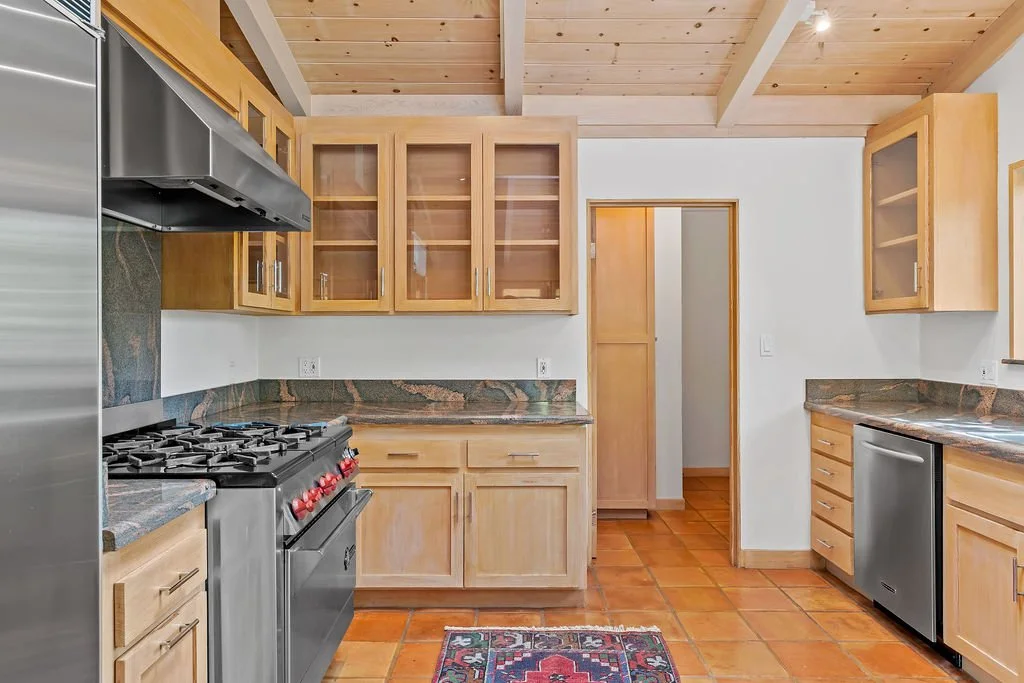 A rustic kitchen with wood cabinets and granite countertops, tiled floor, stainless steel stove, refrigerator, and dishwasher.