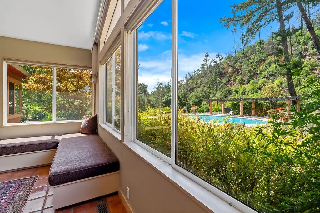 View from inside a room with large windows showing a backyard with a swimming pool, lush green trees, bushes, and a hillside in the background.