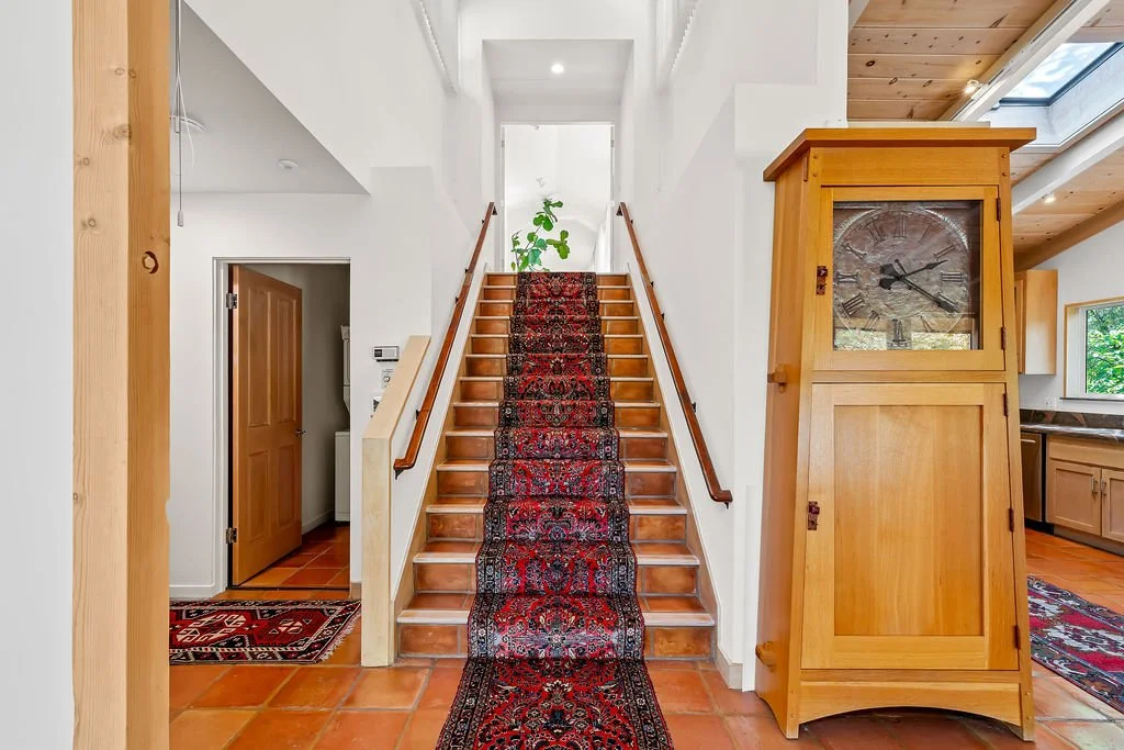 Wooden staircase with a patterned runner, leading to an upper floor with a plant at the top. To the right of the stairs, a tall wooden clock cabinet. The room has terracotta tile flooring, white walls, and wooden accents.