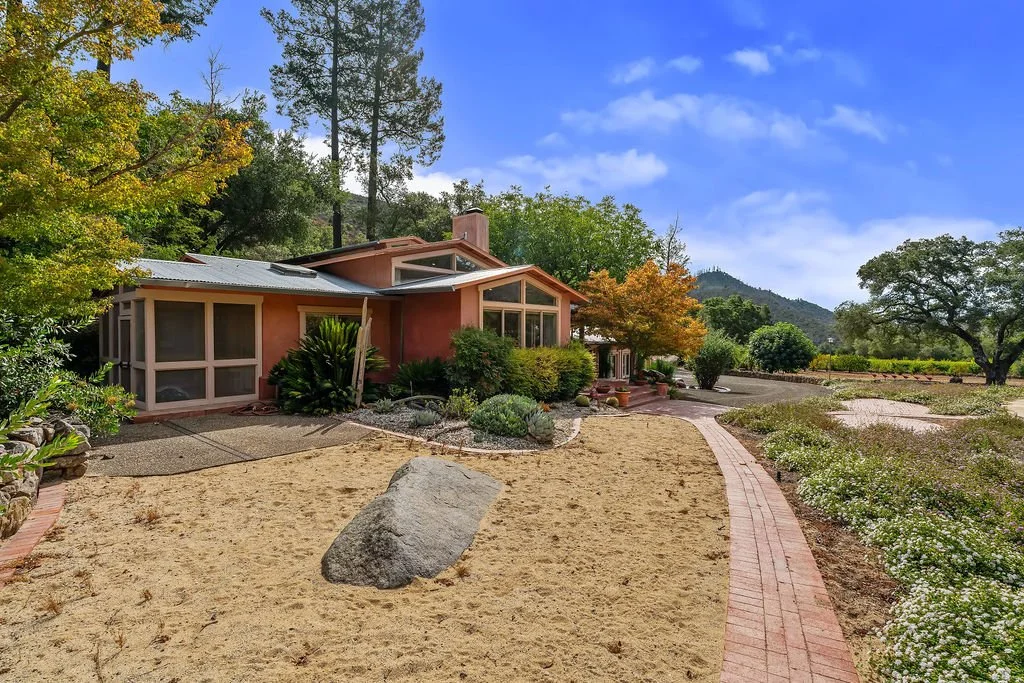 A house with large windows and a garden with trees, bushes, and a sandy pathway, surrounded by mountains under a blue sky with clouds.