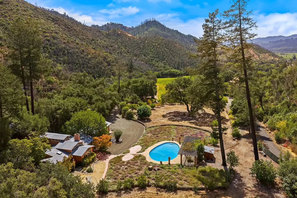 Aerial view of a rural property surrounded by trees and hills, featuring a house, a swimming pool, a pergola, and a dirt road.