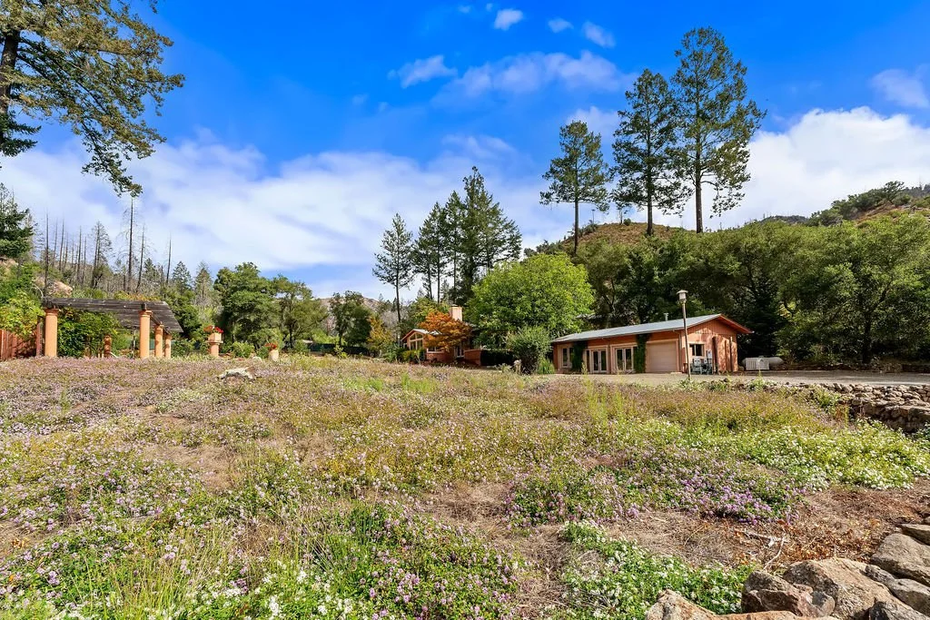 A peaceful landscape with wildflowers in the foreground, a house and a smaller building in the midground, and trees and hills under a bright blue sky with scattered clouds.