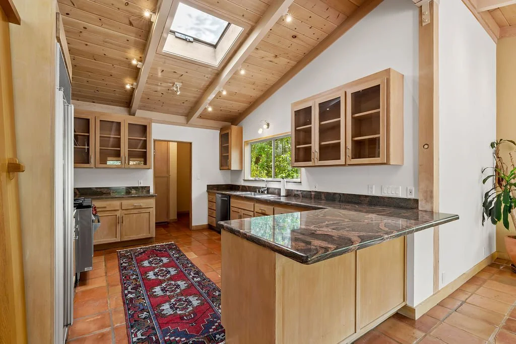 A kitchen with wooden cabinets, a tiled floor, a skylight, and a view through a window of greenery outside.
