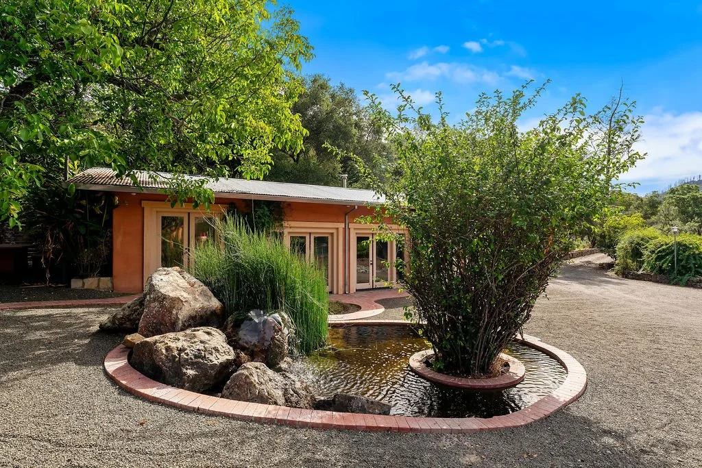 A small house with a metal roof, surrounded by green trees and bushes. In front of the house, there is a decorative water feature with rocks, water, and a bush growing in a circular brick-lined pond, situated on a gravel driveway.