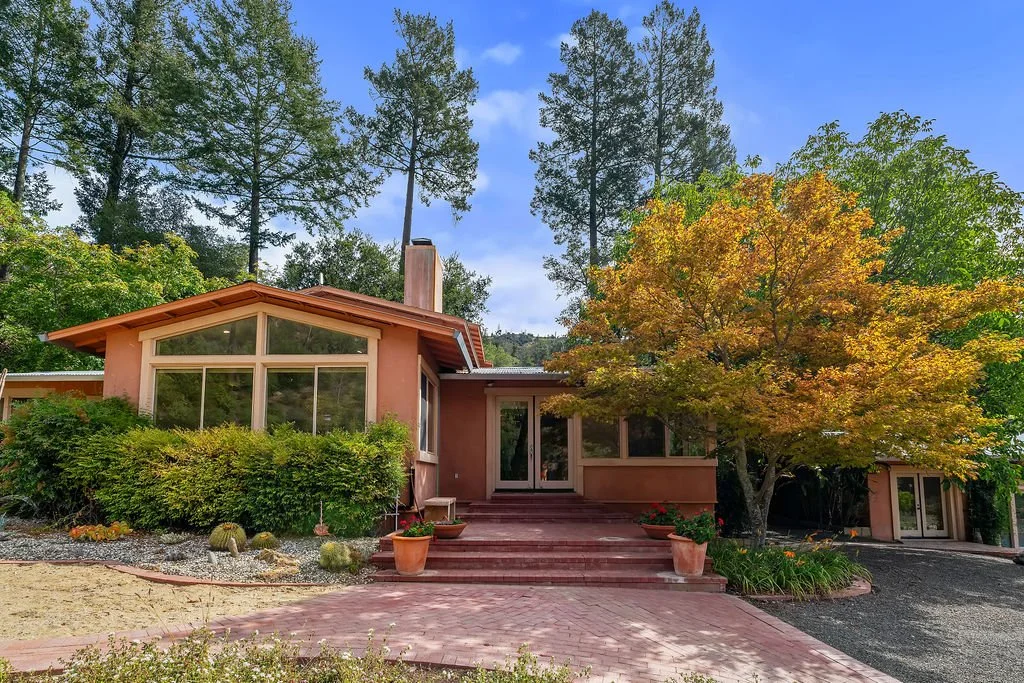 Modern house with large front windows, brick pathway, colorful trees, and lush greenery.