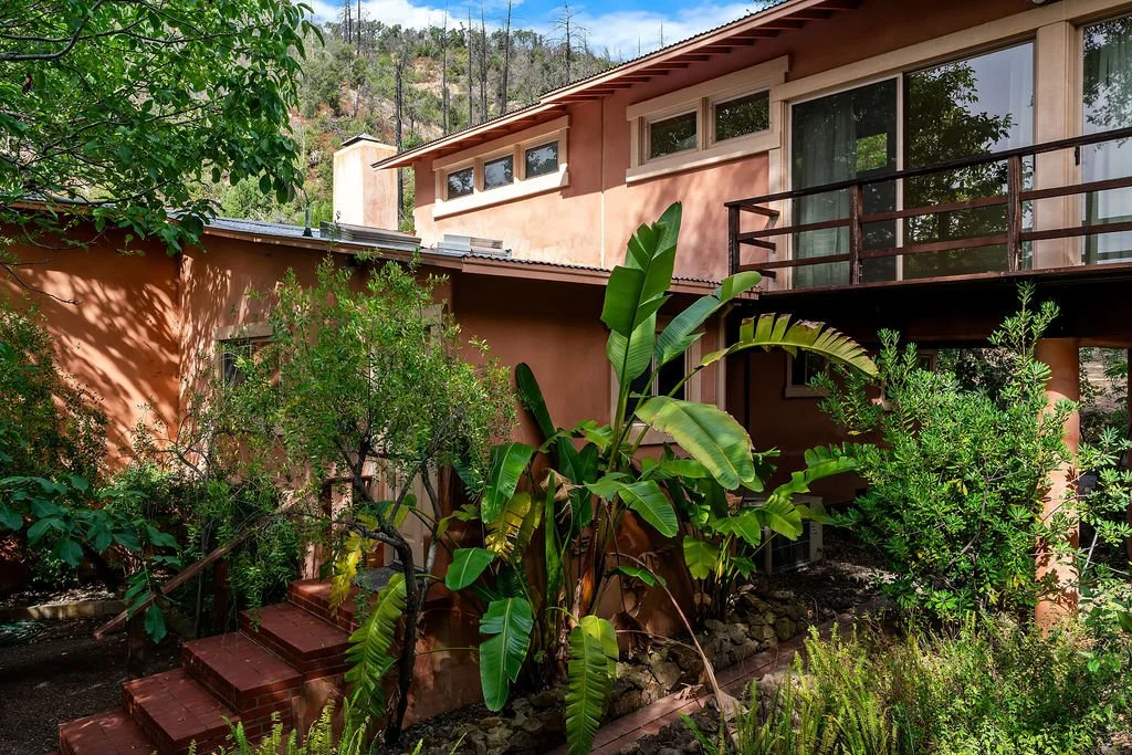A southwestern-style house with a balcony, surrounded by lush green plants, with mountain hills in the background.