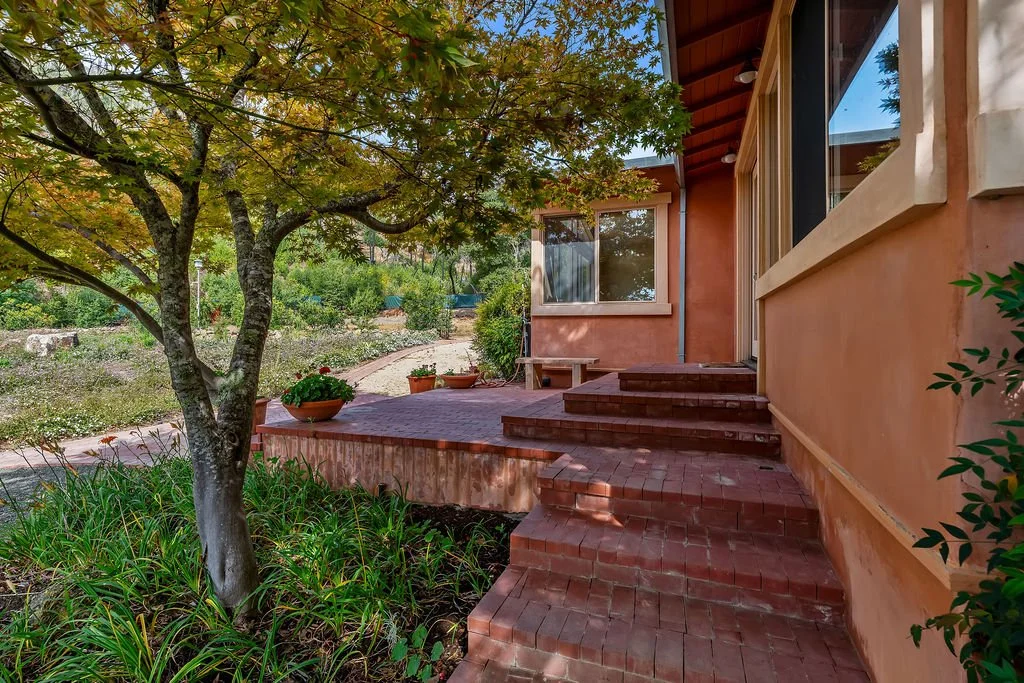 A backyard patio with brick steps leading up to a house with peach-colored stucco walls and large windows. There is a tree with green foliage in the foreground, potted plants on the patio, and a garden area in the background.