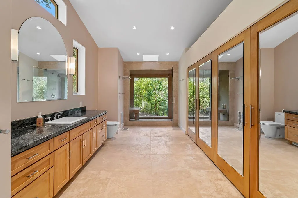 A spacious bathroom with multiple sinks, large mirrors, wooden cabinets, a walk-in shower with glass doors, and a large window overlooking greenery.