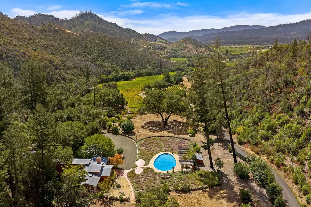 Aerial view of a rural landscape with mountains, green fields, trees, a winding road, a small house, and a swimming pool.