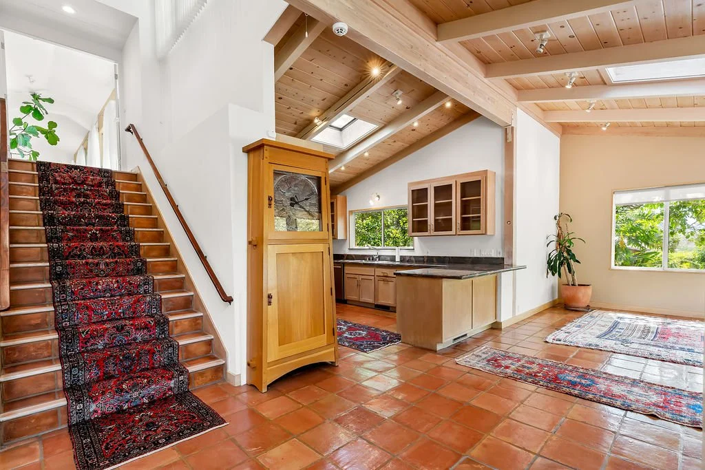 Bright, open-concept living area with terracotta tile flooring, wooden ceiling beams, and large windows with greenery outside. A staircase with a red-patterned runner rug leads upstairs, and the kitchen has wooden cabinets, a black countertop, and a 