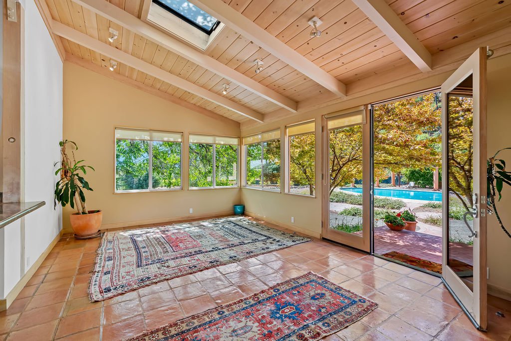 Empty sunroom with wooden ceiling and terracotta tile floor, decorated with rugs, potted plants, large windows, and a door leading outside to a patio and swimming pool.