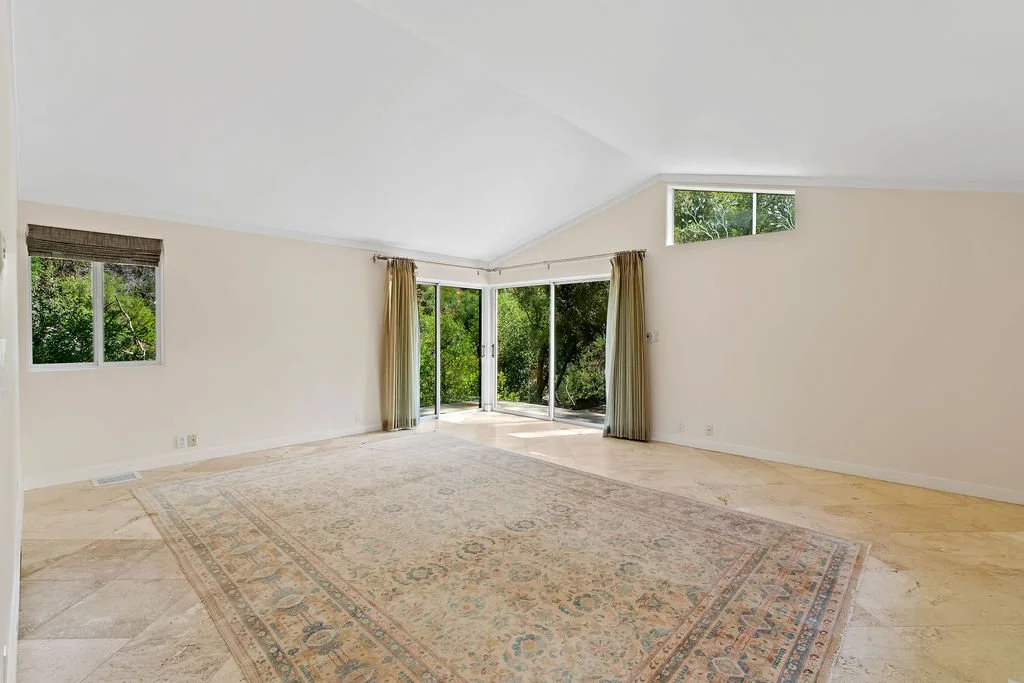 Empty living room with large windows, sliding glass door with curtains, beige walls, a large patterned area rug, and tile flooring.